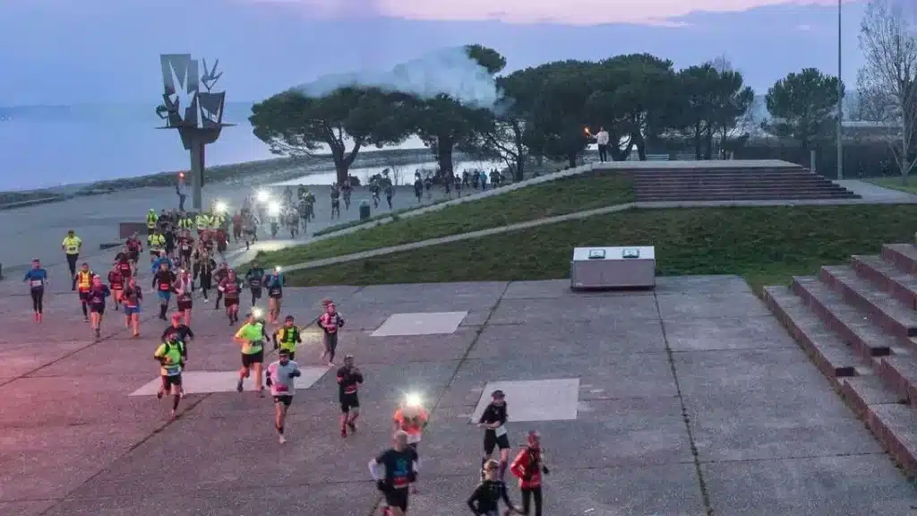 Une course nocturne est en cours, avec des participants vêtus de tenues de sport et de gilets réfléchissants. Ils courent le long d'un sentier pavé près d'un plan d'eau, avec un feu d'artifice illuminant le crépuscule. À l'arrière-plan, le ciel est teinté de nuances de rose et de bleu au crépuscule. Des arbres et des sculptures modernes ornent le paysage environnant, ajoutant à l'atmosphère festive et dynamique de l'événement
