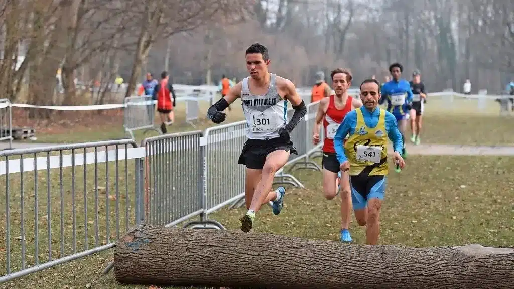 Coureurs en compétition sautant par-dessus un tronc d'arbre lors d'un cross-country, illustrant le dynamisme et les défis physiques du sport