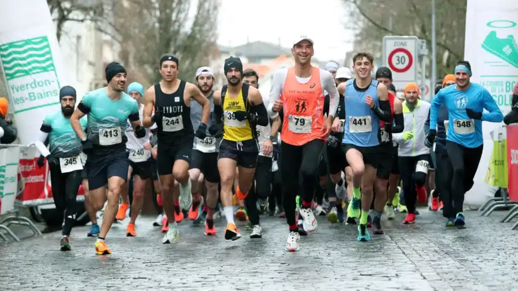 Un peloton de coureurs débute une course sur des pavés humides, probablement l'événement "La Course de la Bonne" à Yverdon-les-Bains, au vu des dossards et des bannières. Le dynamisme est palpable, avec des expressions concentrées et souriantes malgré le temps gris, ce qui reflète bien l'esprit de camaraderie et de détermination du running communautaire.