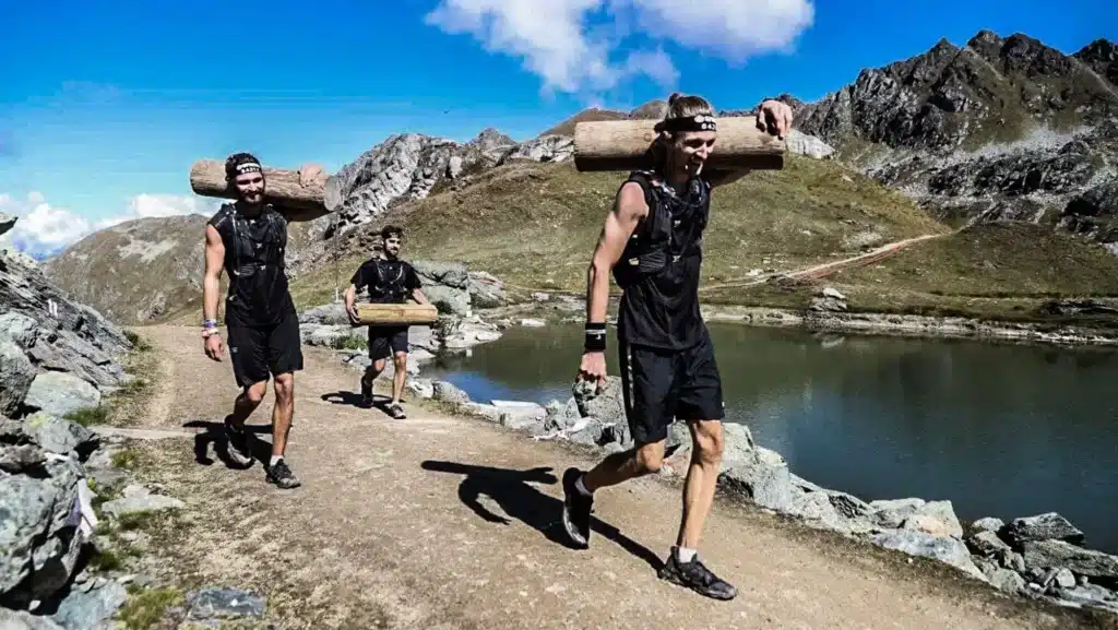 Cette image montre trois individus participant à une activité de plein air, probablement une course de type spartiate ou trail, au vu de leurs tenues sportives et du fait qu'ils portent des rondins de bois sur leurs épaules. Ils sont en mouvement sur un sentier de montagne rocheux. À l'arrière-plan, on peut voir un lac de montagne tranquille et des crêtes montagneuses. Le paysage suggère que l'événement se déroule dans une région alpine, et la luminosité indique qu'il s'agit d'une journée ensoleillée. Les participants sont concentrés sur leur effort, ce qui témoigne de la nature exigeante de l'épreuve. Course Spartan Verbier