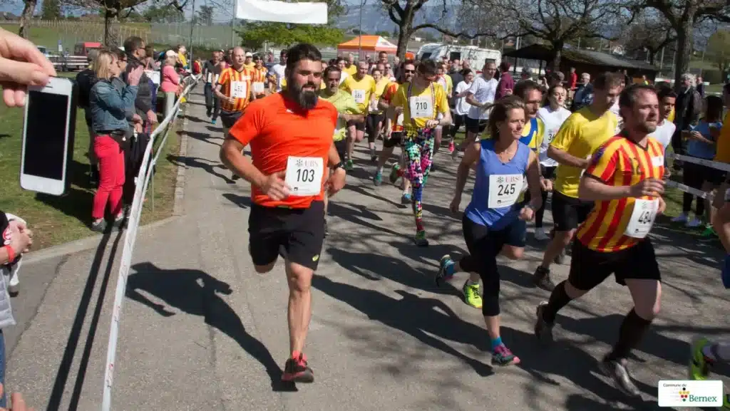 Des coureurs en pleine action lors d'une course pédestre, avec des spectateurs les encourageant sur le côté. Les participants, numérotés et en tenue de sport, courent sur une route goudronnée par une journée ensoleillée. L'ambiance est dynamique et festive. Course de Bernex.