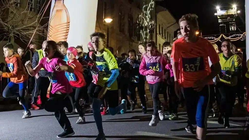 Jeunes coureurs enthousiastes prenant un départ rapide lors d'une course nocturne à Avenches, éclairés par les lumières de la ville et encouragés par les spectateurs, capturant l'énergie et l'excitation de l'événement sportif