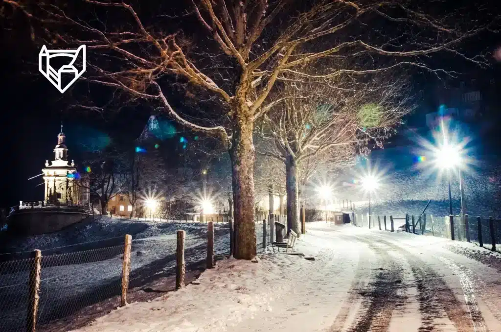 Une nuit hivernale paisible se dépeint ici, avec un chemin enneigé menant à une chapelle éclairée, tandis que les lampadaires répandent une lueur chaleureuse sous le ciel nocturne et les arbres dénudés se dressent en sentinelles silencieuses.