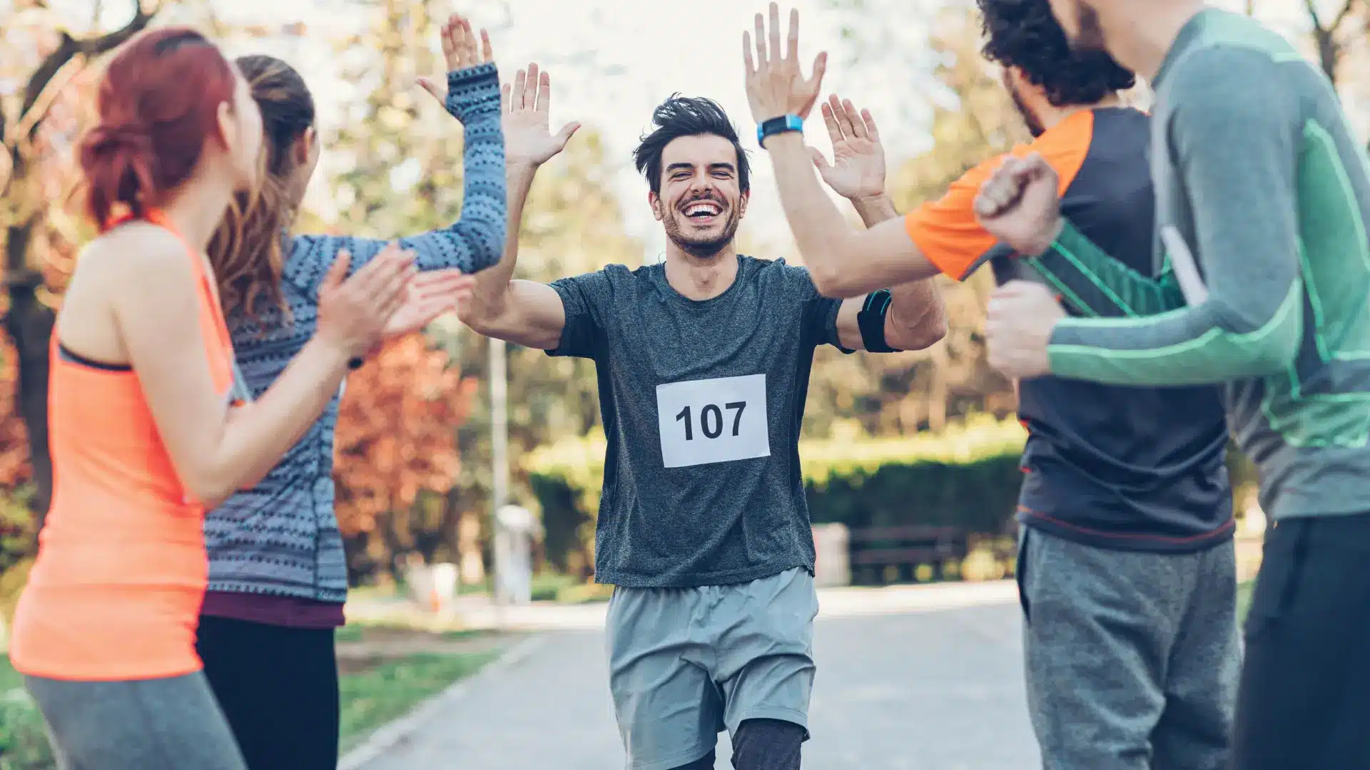 L'image montre un moment joyeux lors d'un événement de course à pied, probablement un coureur masculin franchissant la ligne d'arrivée et étant accueilli par les acclamations et les high-fives des spectateurs et autres coureurs. Il porte un dossard avec le numéro 107 et a un large sourire, ce qui indique qu'il est très content de sa performance.